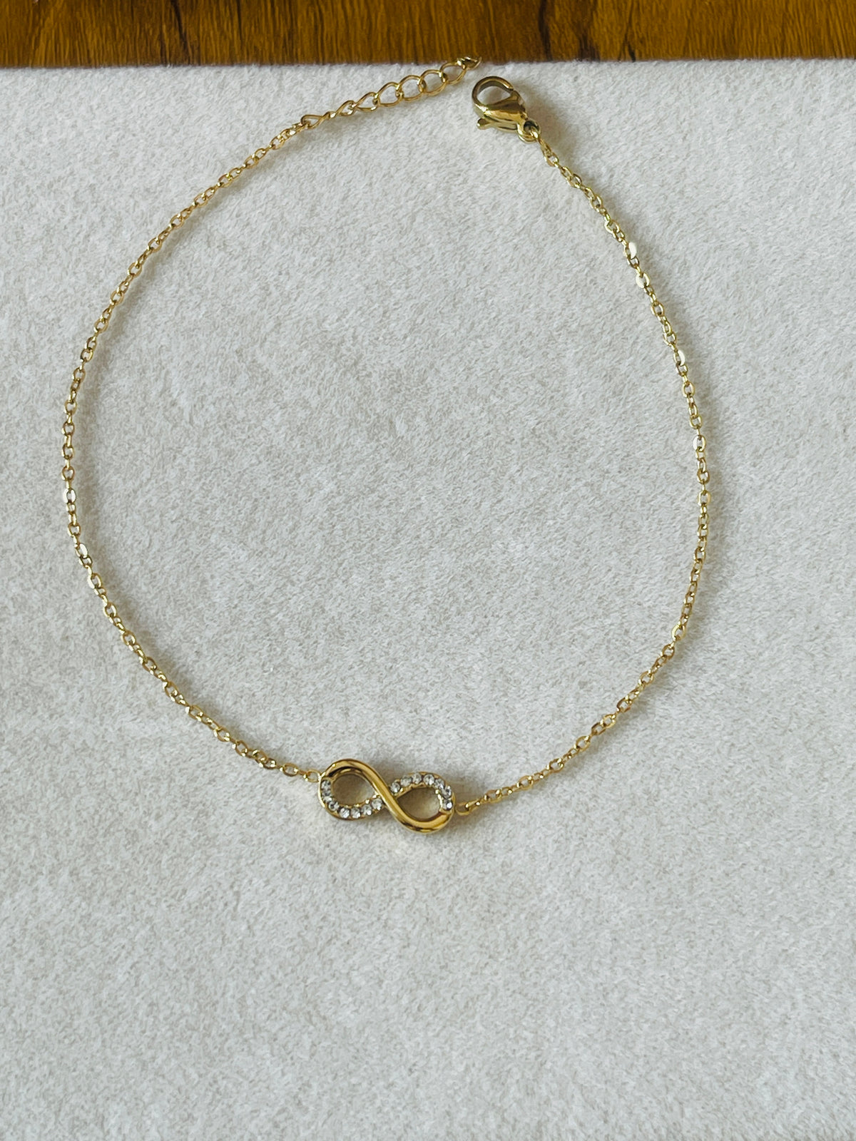 A delicate gold chain bracelet with an infinity symbol charm set with small clear stones, displayed on a textured white surface.