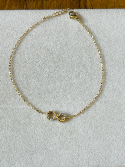 A delicate gold chain bracelet with an infinity symbol charm set with small clear stones, displayed on a textured white surface.