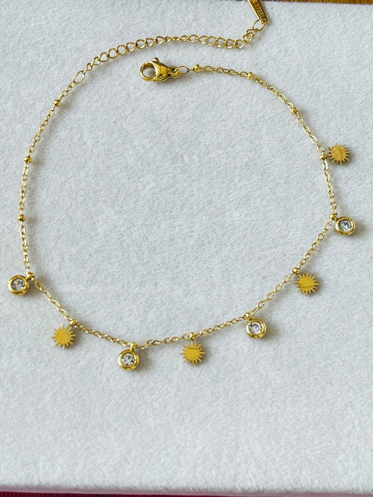 A gold chain anklet featuring alternating sunburst charms and clear crystal bezel settings, displayed on a textured white background.