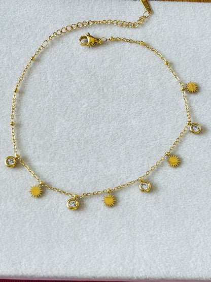A gold chain anklet featuring alternating sunburst charms and clear crystal bezel settings, displayed on a textured white background.