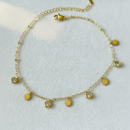 A delicate gold chain necklace with small sunburst charms and three clear crystal bezel settings, displayed on a textured light gray background.