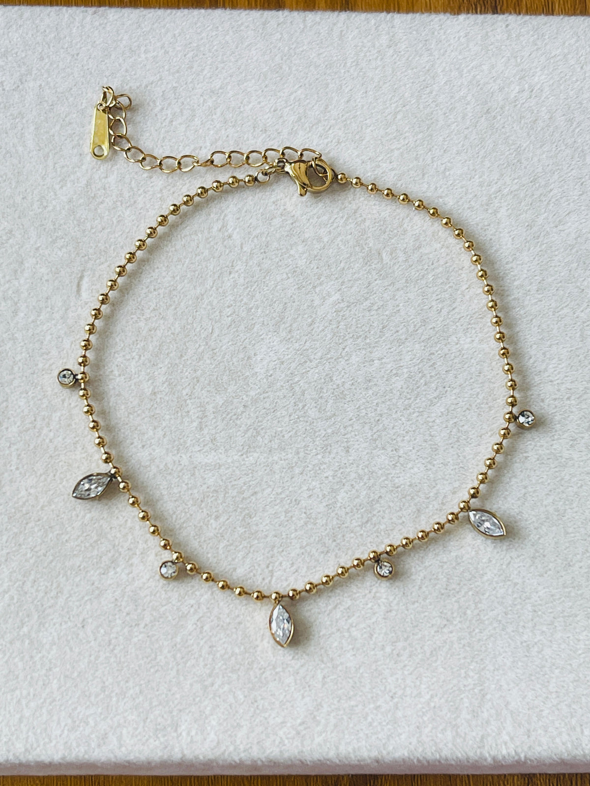A gold beaded anklet with small clear marquise and round crystals, displayed on a white textured background.