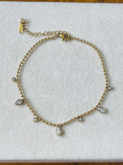 A gold beaded anklet with small clear marquise and round crystals, displayed on a white textured background.