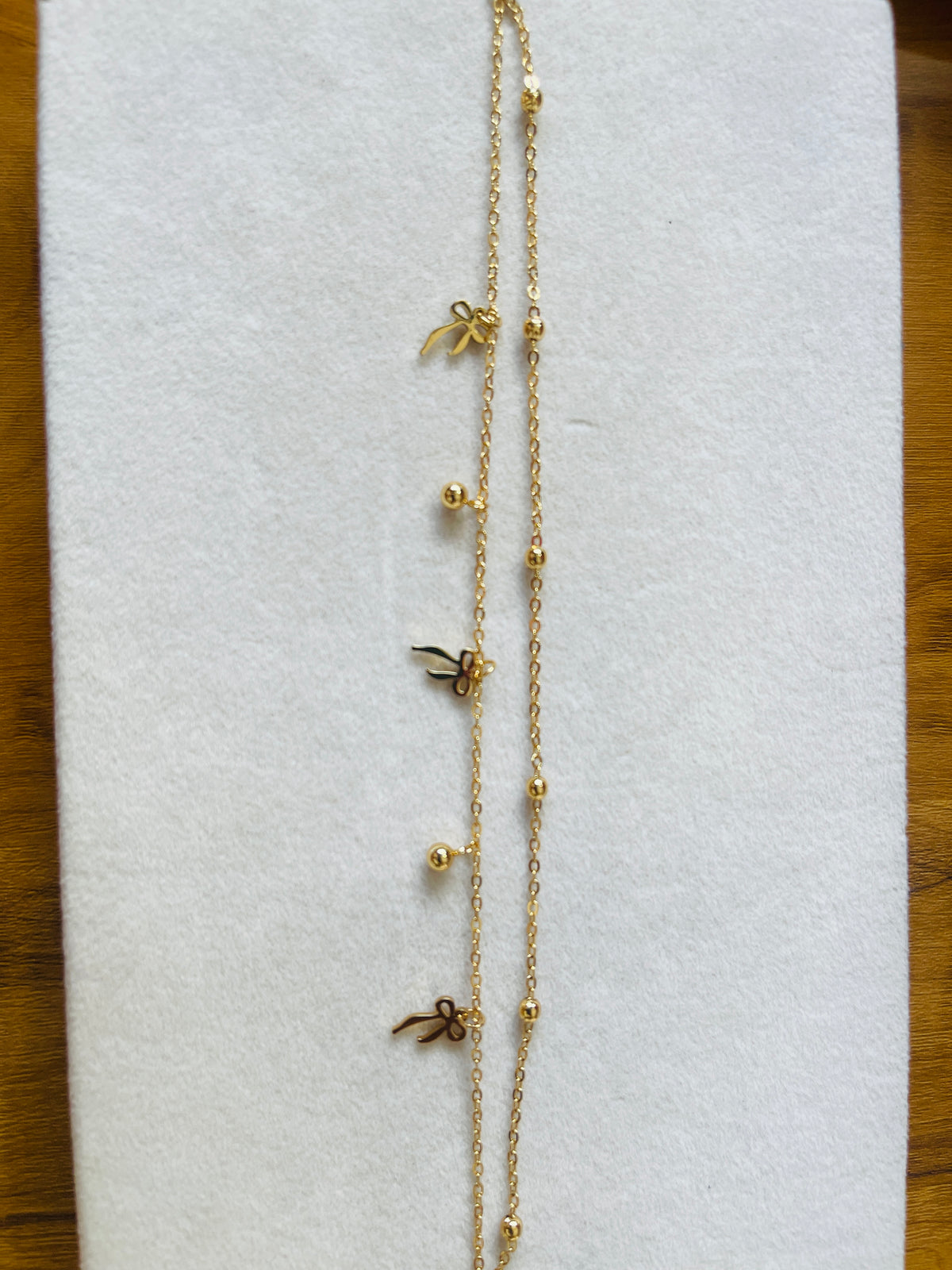 A delicate gold chain necklace laid flat on a white textured display card, featuring small gold bead accents and three tiny bow-shaped charms.