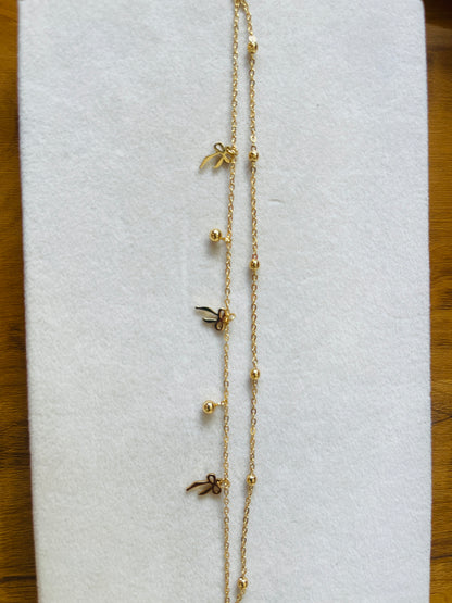A delicate gold chain necklace laid flat on a white textured display card, featuring small gold bead accents and three tiny bow-shaped charms.