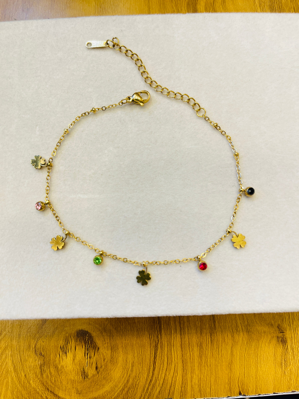 A gold chain anklet with multiple dangling charms, including four-leaf clovers, small gold leaves, and tiny colored beads, displayed on a white textured surface.