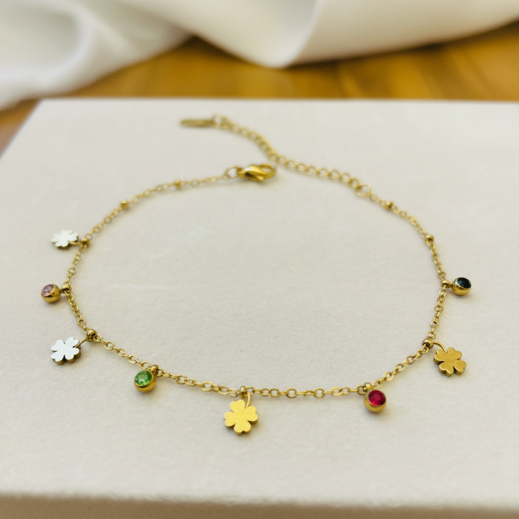 A delicate gold chain anklet with dangling charms, including four gold clover shapes and several small round gemstone charms in white, green, pink, and black, displayed on a light cream jewelry box.