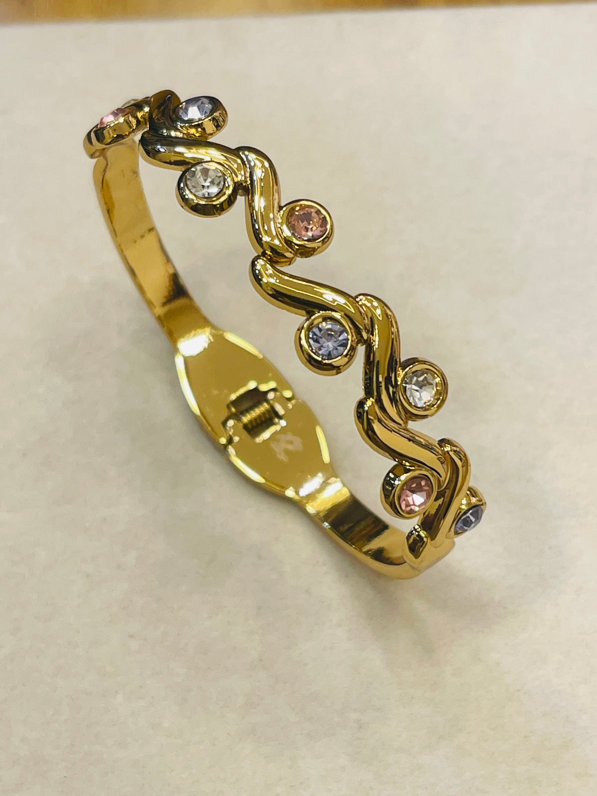 A gold-plated bangle bracelet with a wavy, vine-like design adorned with small clear and pink gemstones, displayed on a light beige textured surface.