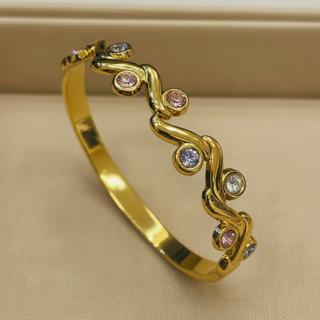 A polished gold bangle bracelet with a wavy vine-like design set with small round gemstones in pink, clear, and light purple, displayed on a beige textured surface.
