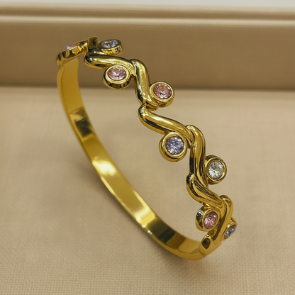 A polished gold bangle bracelet with a wavy vine-like design set with small round gemstones in pink, clear, and light purple, displayed on a beige textured surface.
