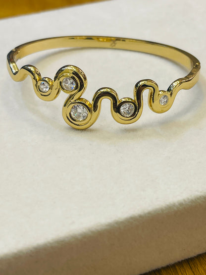 A gold wave-shaped cuff bracelet with three round clear gemstones, displayed on a cream-colored textured surface.