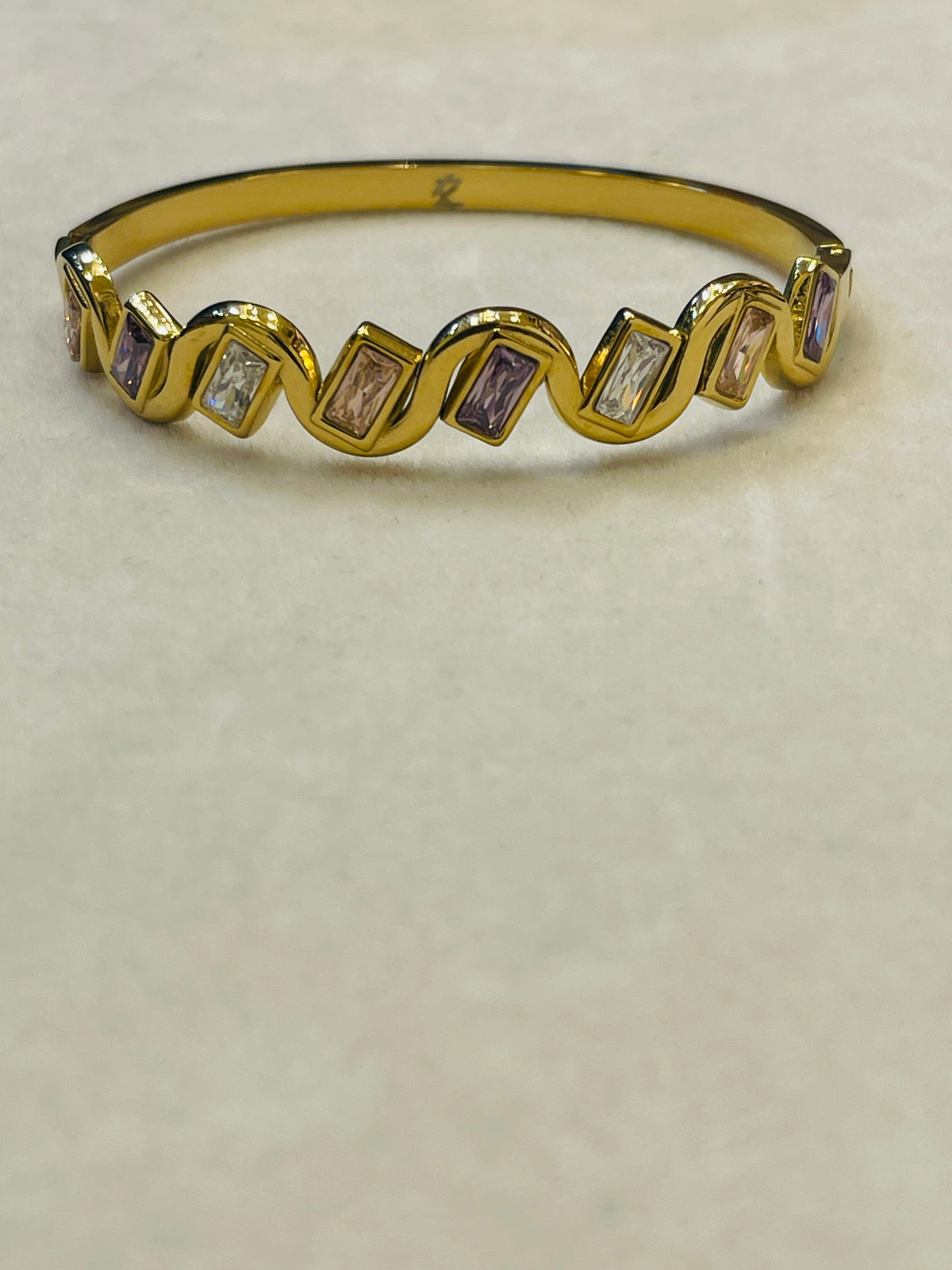 A gold band ring featuring a wavy zigzag pattern with alternating clear baguette-cut stones and lavender baguette-cut stones, displayed on a neutral beige surface.