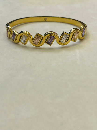 A gold band ring featuring a wavy zigzag pattern with alternating clear baguette-cut stones and lavender baguette-cut stones, displayed on a neutral beige surface.