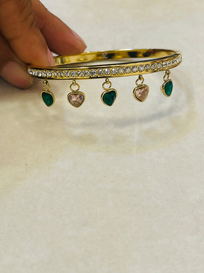 A gold-plated hinged bangle bracelet with a row of clear crystals and five dangling heart charms (four green, one pink), held by a hand against a light beige background.
