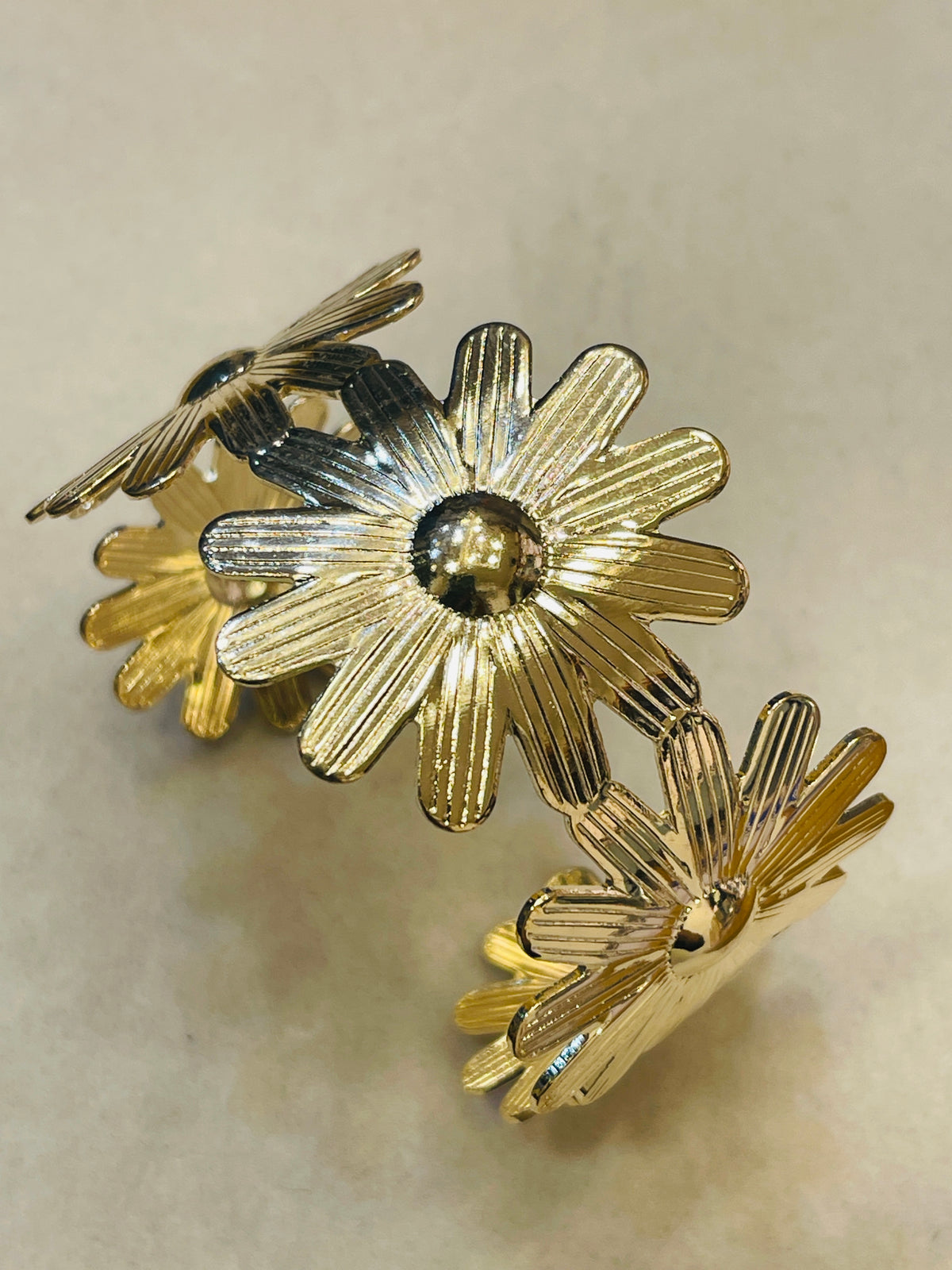A gold-tone metal cuff bracelet featuring a series of textured daisy flower motifs with polished centers, displayed on a neutral beige background.