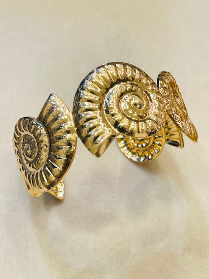 A pair of gold-colored spiral shell earrings shaped like ammonites, displayed on a neutral beige background.