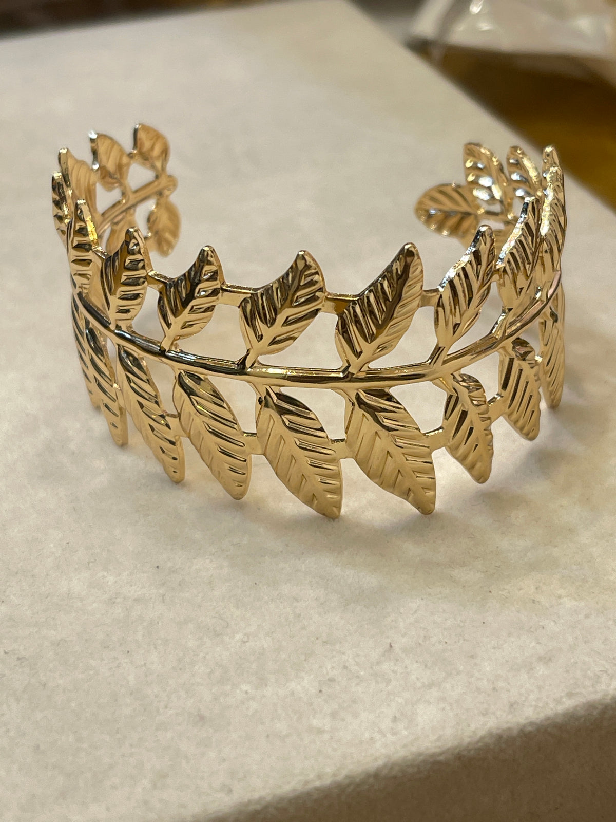 A gold leaf-patterned cuff bracelet displayed on a textured beige surface.