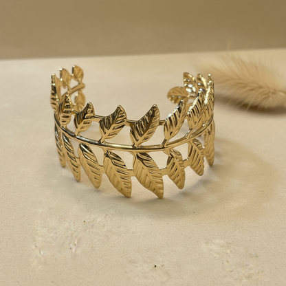 A gold leaf-patterned cuff bracelet displayed on a textured beige surface with a soft beige background.