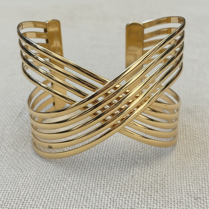 A gold-toned metal cuff bracelet with multiple parallel and crossed bands, photographed on a textured off-white fabric background.