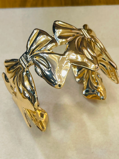 A chunky metallic cuff bracelet featuring a large bow design, finished in a shiny gold-tone metal with reflective silver-toned highlights, displayed on a light beige surface.