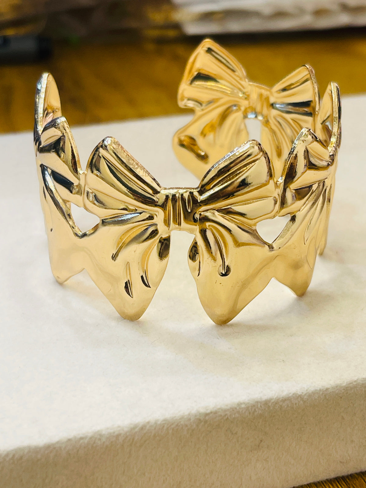 A shiny gold bow-shaped cuff bracelet displayed on a cream-colored textured surface.