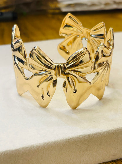 A shiny gold bow-shaped cuff bracelet displayed on a cream-colored textured surface.