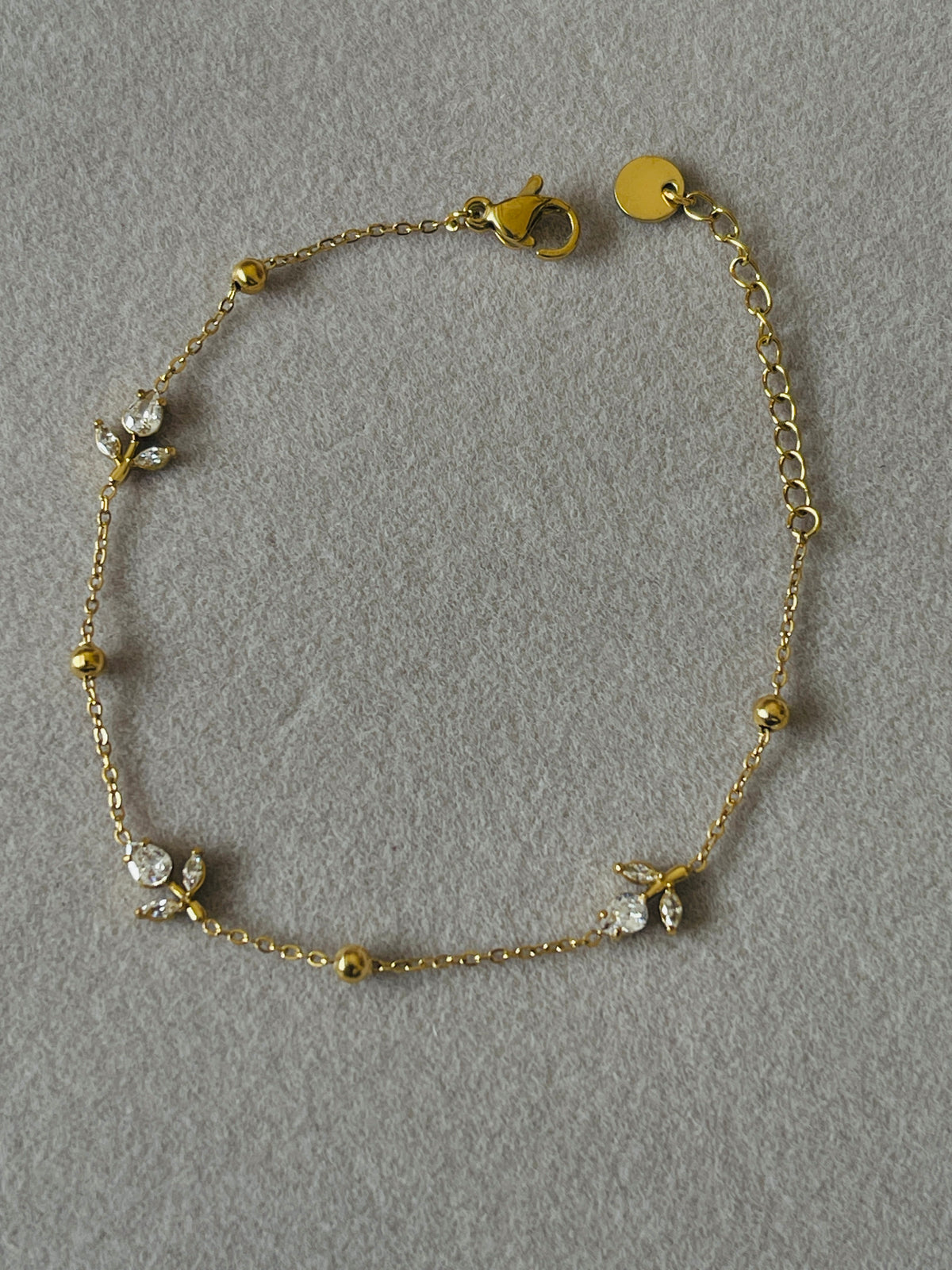 A delicate gold chain bracelet with small bead accents and three floral clusters set with clear rhinestones, displayed on a textured gray background.