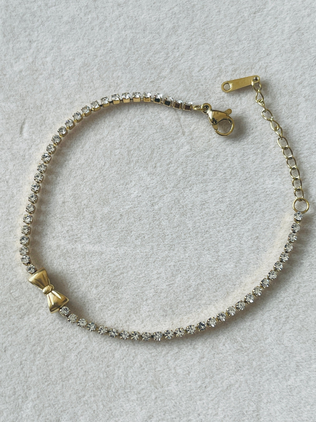 A delicate gold chain bracelet featuring a row of clear rhinestones, a small gold bow charm, and a lobster clasp closure, displayed on a textured light gray background.