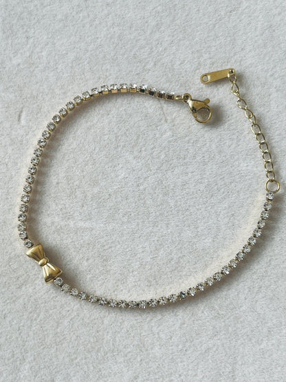 A delicate gold chain bracelet featuring a row of clear rhinestones, a small gold bow charm, and a lobster clasp closure, displayed on a textured light gray background.