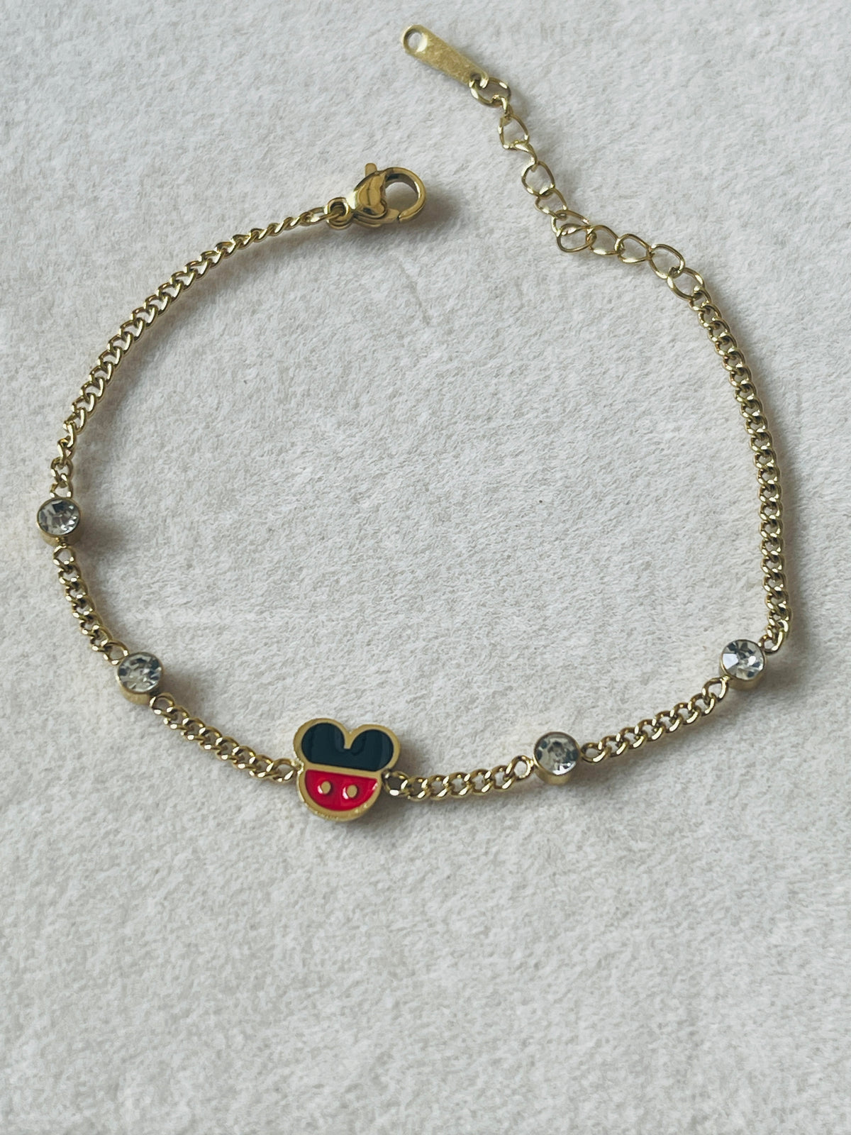 A gold chain bracelet featuring a Mickey Mouse head charm with red and blue enamel, accented by clear rhinestones on a textured white background.