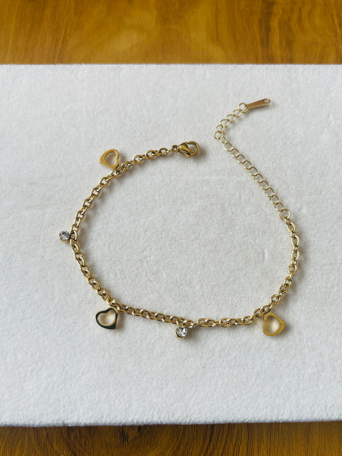 A gold chain bracelet with heart charms, displayed on a white textured foam pad over a wooden surface.