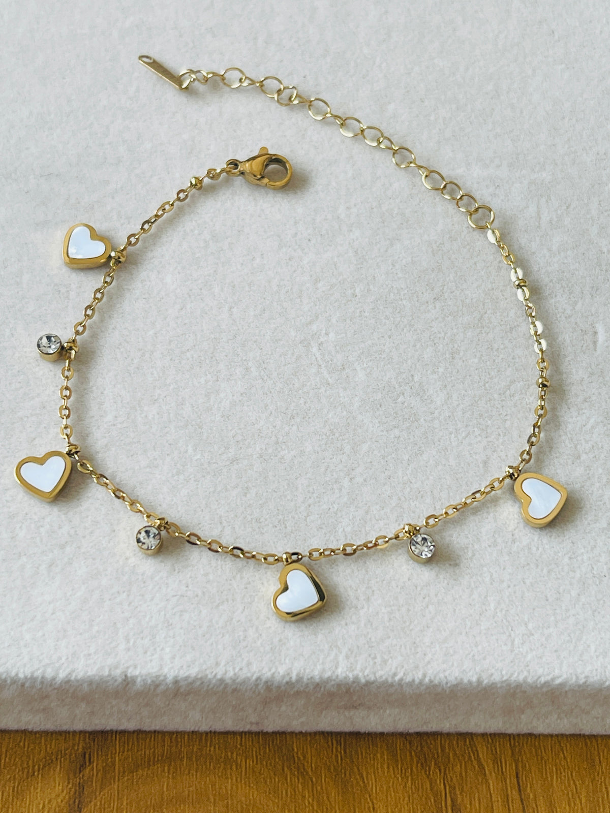 A gold chain bracelet featuring five heart charms, three with white inlay and two with clear rhinestones, displayed on a textured white surface.