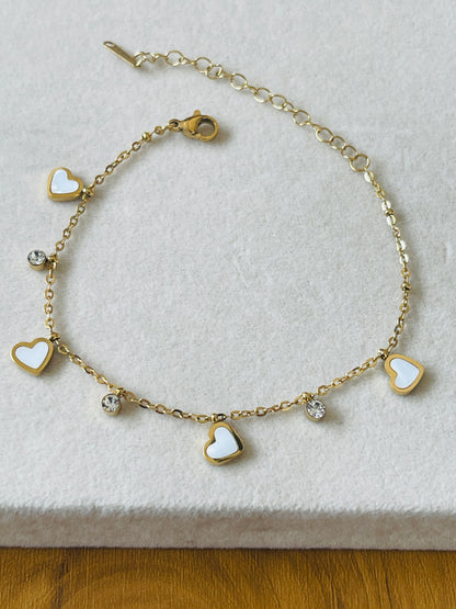 A gold chain bracelet featuring five heart charms, three with white inlay and two with clear rhinestones, displayed on a textured white surface.
