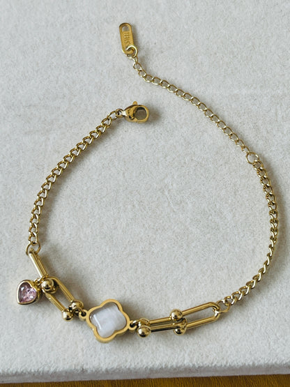 A gold chain bracelet with a four-leaf clover charm, a pink heart charm, and a white oval charm, displayed on a textured white surface.