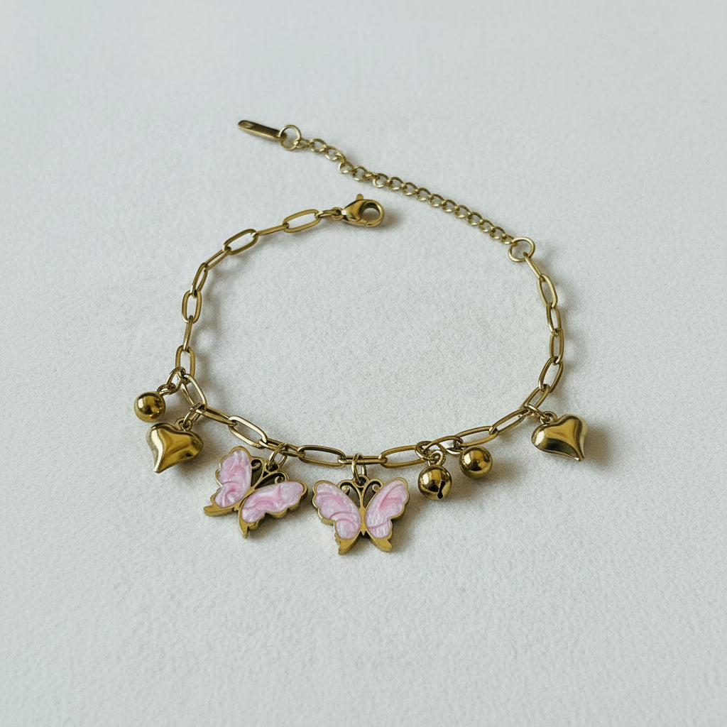 A gold chain bracelet with paperclip links, small gold beads, and three charms: a pink and gold butterfly, two gold hearts, and a gold heart with a pink enamel center, displayed on a textured white surface.