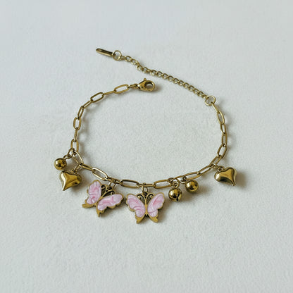 A gold chain bracelet with paperclip links, small gold beads, and three charms: a pink and gold butterfly, two gold hearts, and a gold heart with a pink enamel center, displayed on a textured white surface.
