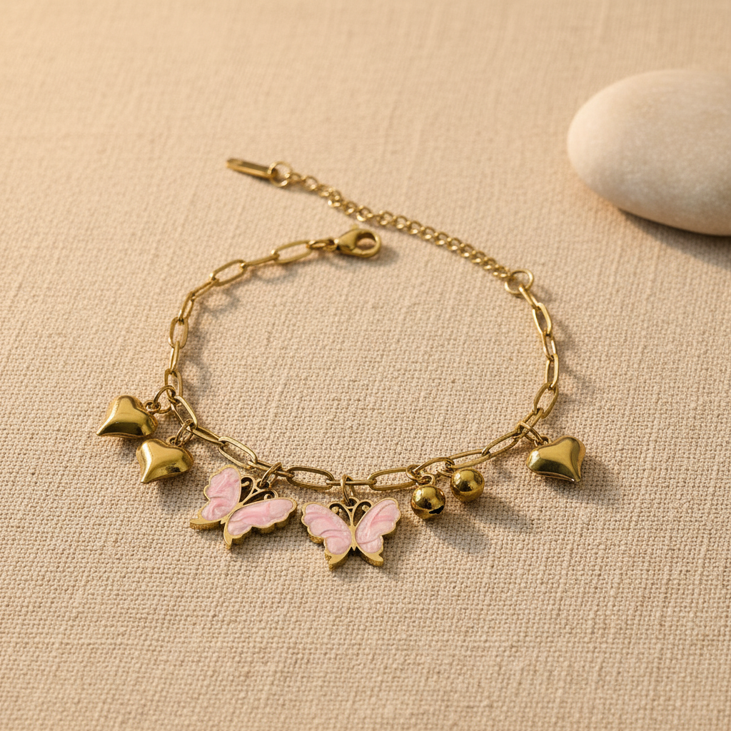 A gold-tone chain bracelet with dangling charms, including two pink-and-gold butterflies, two gold hearts, and two small gold beads, displayed on a textured beige fabric surface with a smooth white stone prop.