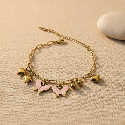 A gold-tone chain bracelet with dangling charms, including two pink-and-gold butterflies, two gold hearts, and two small gold beads, displayed on a textured beige fabric surface with a smooth white stone prop.