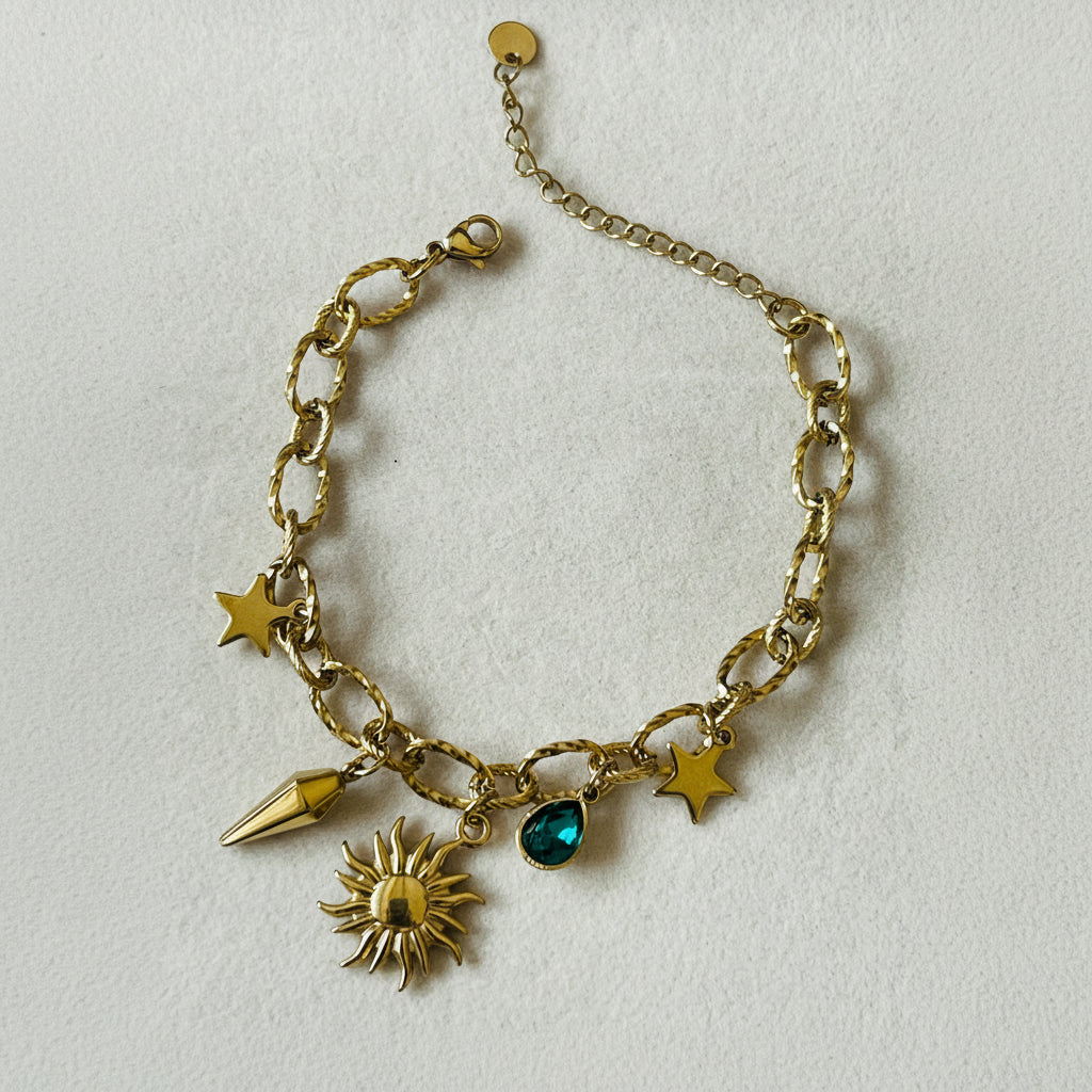A gold-tone charm bracelet with a link chain, featuring sun, star, and spearhead charms, plus a small green teardrop gemstone, displayed on a white textured surface.