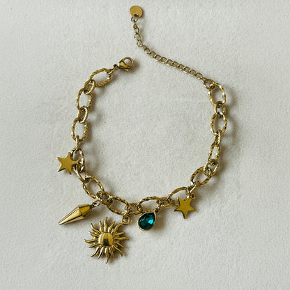 A gold-tone charm bracelet with a link chain, featuring sun, star, and spearhead charms, plus a small green teardrop gemstone, displayed on a white textured surface.
