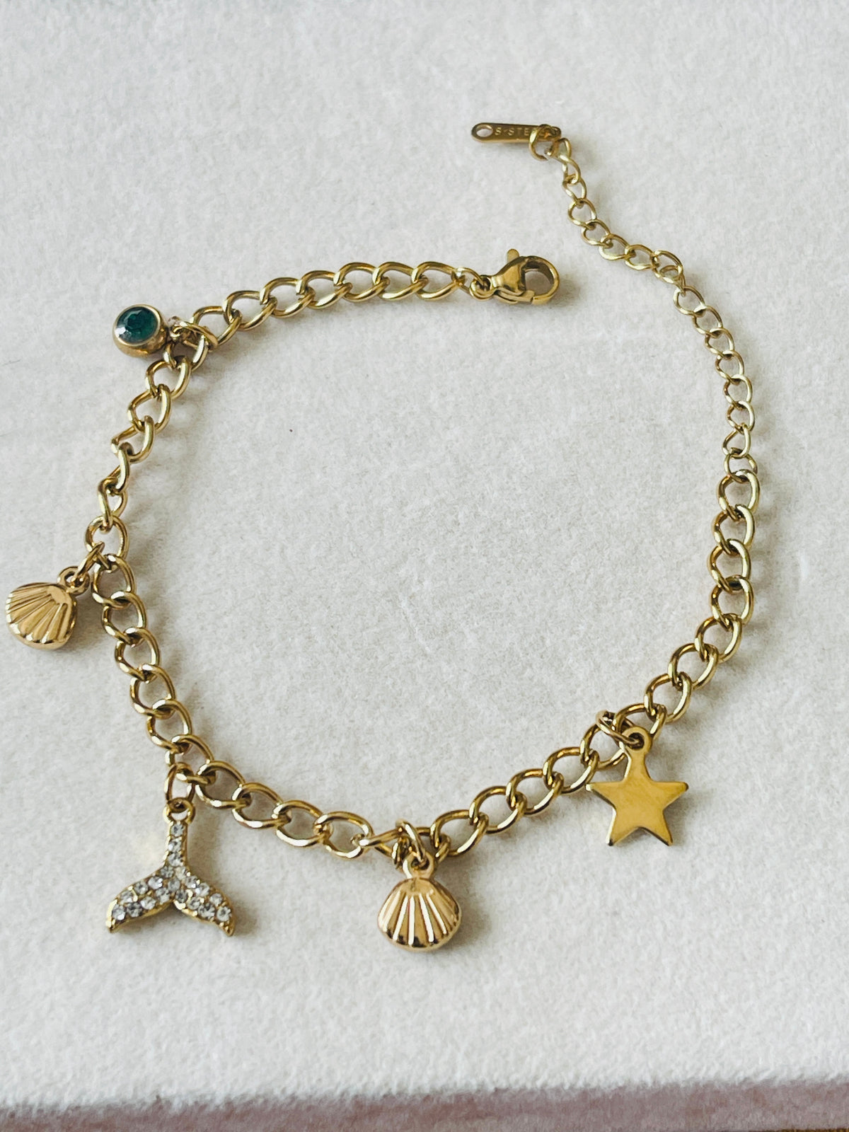 A gold-tone chain bracelet with dangling charms including a starfish, seashells, a mermaid tail, and a small green stone accent, displayed on a textured white surface.
