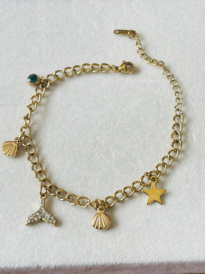 A gold-tone chain bracelet with dangling charms including a starfish, seashells, a mermaid tail, and a small green stone accent, displayed on a textured white surface.