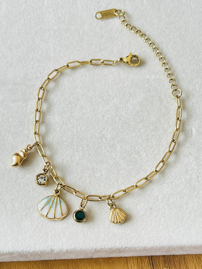 A gold chain anklet with shell and seashell charms, displayed on a textured white fabric background.