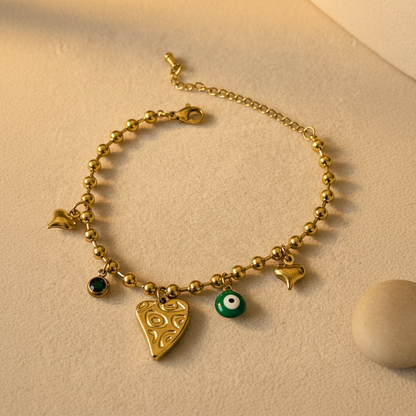 A gold beaded charm bracelet featuring a large gold heart pendant, a small green evil eye charm, a dark green round charm, and three small gold heart charms, displayed on a textured beige surface.
