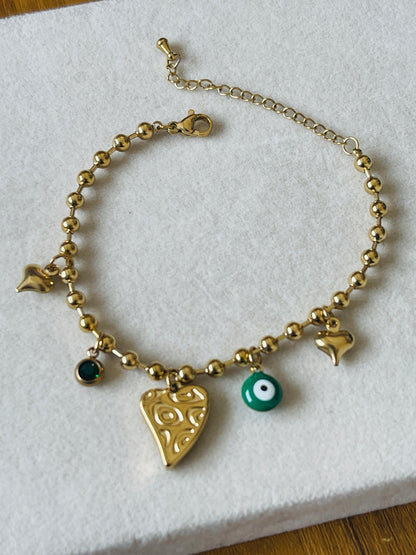 A gold beaded charm bracelet displayed on a white textured surface. The bracelet features multiple charms, including a large gold heart with a carved pattern, a small green evil eye bead, a dark green round bead, a small gold star, and two smaller gold heart charms, with an adjustable gold chain and clasp.