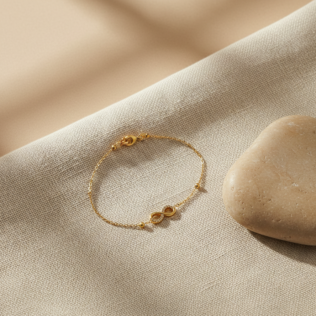 A delicate gold chain bracelet with a small infinity symbol charm, resting on a textured beige linen fabric next to a smooth stone.