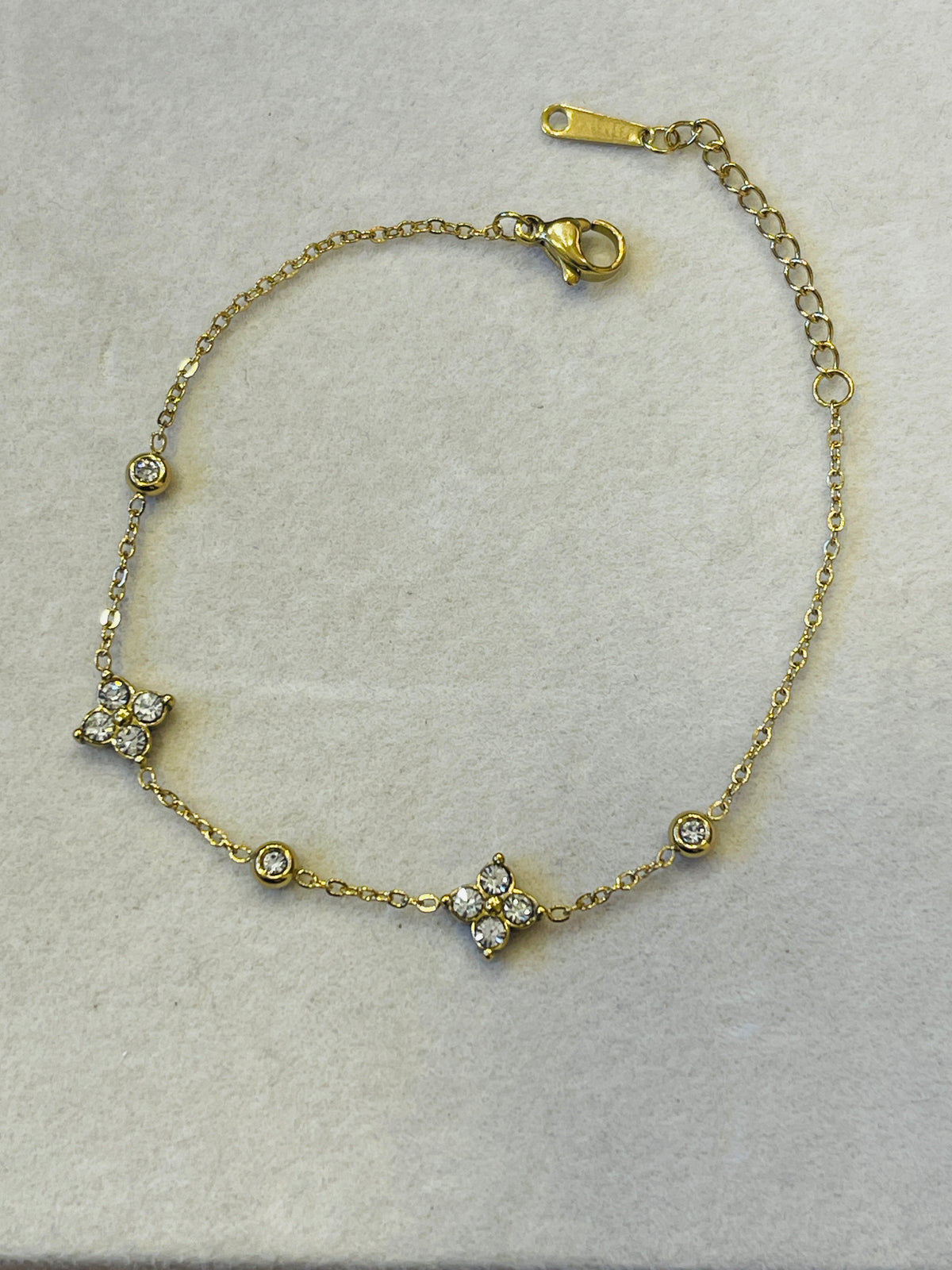 A delicate gold chain bracelet with three floral clusters set with clear stones, displayed on a textured light gray surface.