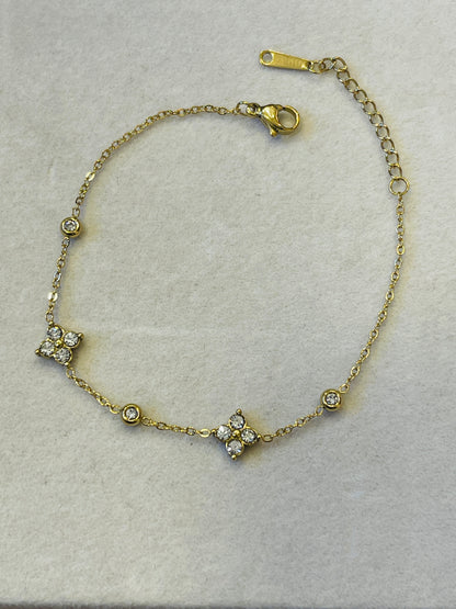 A delicate gold chain bracelet with three floral clusters set with clear stones, displayed on a textured light gray surface.