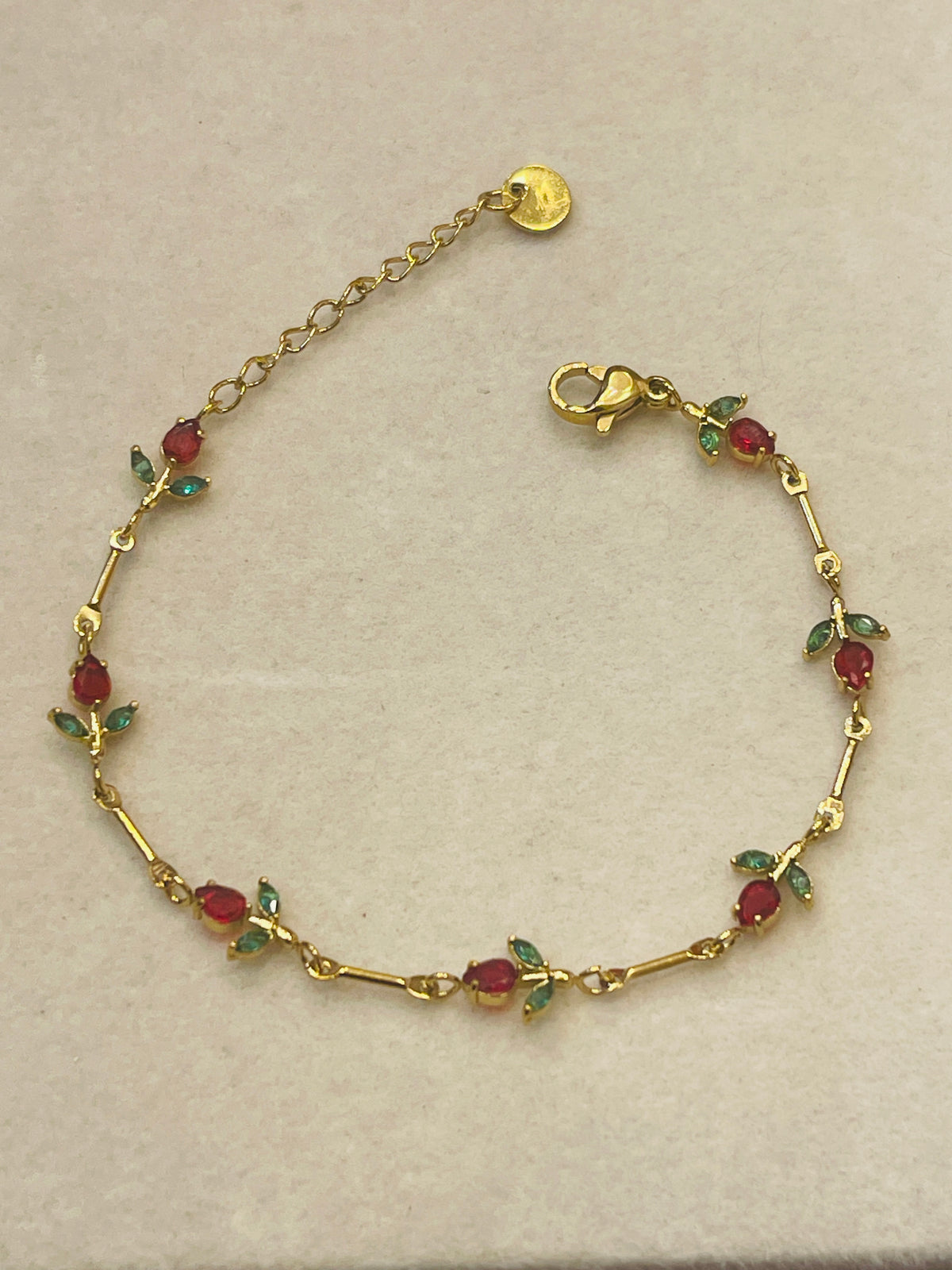 A vintage yellow gold bracelet featuring small oval red gemstones and green leaf-shaped accents, displayed on a neutral beige background.