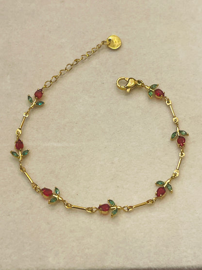 A vintage yellow gold bracelet featuring small oval red gemstones and green leaf-shaped accents, displayed on a neutral beige background.